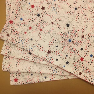 Peut inclure: Quatre serviettes en tissu blanc avec un motif d'&eacute;toiles rouges, bleues et noires. Les serviettes sont pli&eacute;es et empil&eacute;es les unes sur les autres.