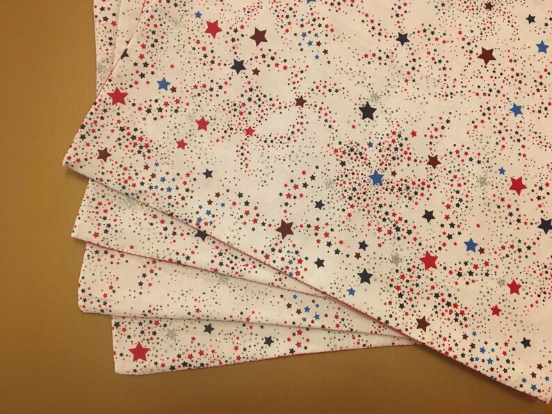 Peut inclure: Quatre serviettes en tissu blanc avec un motif d'&eacute;toiles rouges, bleues et noires. Les serviettes sont pli&eacute;es et empil&eacute;es les unes sur les autres.