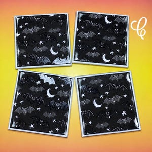 May include: Four square coasters featuring a repeating pattern of black bats, white crescent moons, and white stars on a black background. Each coaster has a glossy finish and a white border, arranged in a two-by-two grid formation.