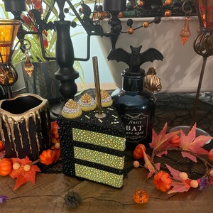 May include: A black and gold cake slice with rhinestones, a black candle holder with a bat design, and a black bottle with the text "finest bat ave aged" on a wooden table with autumn decorations.