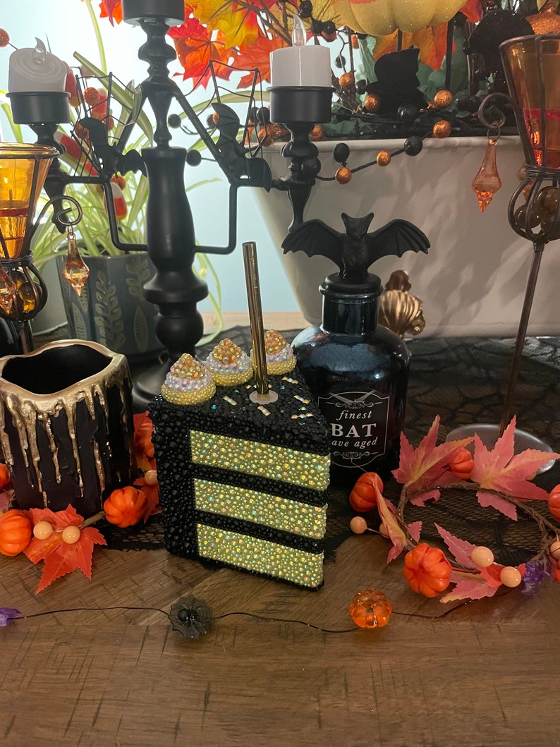 May include: A black and gold cake slice with rhinestones, a black candle holder with a bat design, and a black bottle with the text "finest bat ave aged" on a wooden table with autumn decorations.