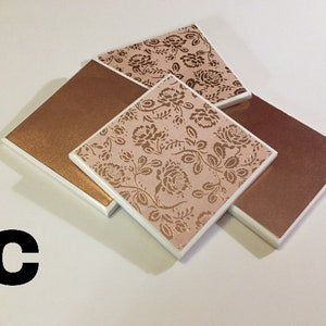 Puede incluir: Conjunto de cuatro posavasos con un dise&ntilde;o floral con detalles dorados sobre un fondo rosa. Dos de los posavasos son de cobre macizo.