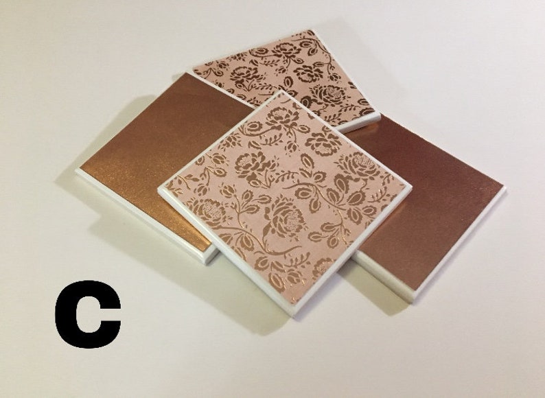 Puede incluir: Conjunto de cuatro posavasos con un dise&ntilde;o floral con detalles dorados sobre un fondo rosa. Dos de los posavasos son de cobre macizo.