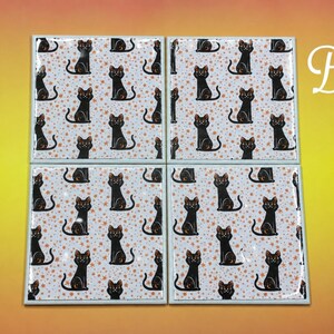 May include: Four square ceramic coasters featuring a repeating pattern of black cats on a white background with small orange stars. The letter B is printed in white in the upper right corner. Each coaster has a glossy finish and a light grey border.