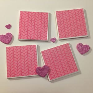 May include: Four pink and white ceramic coasters with a pink leaf pattern. The coasters are arranged in a square shape with a pink glitter heart on each corner.