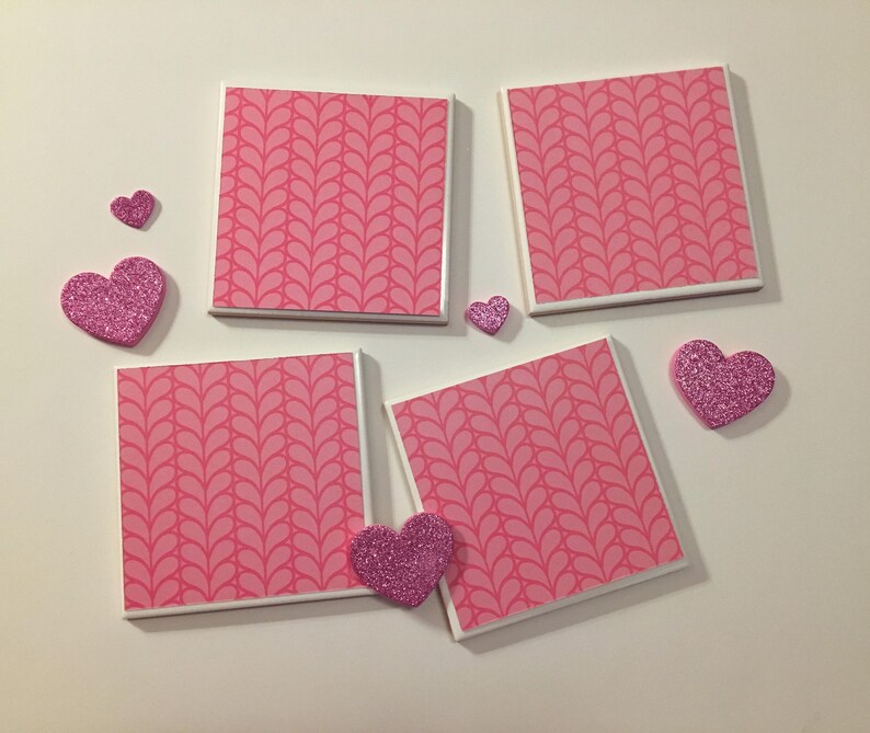 May include: Four pink and white ceramic coasters with a pink leaf pattern. The coasters are arranged in a square shape with a pink glitter heart on each corner.