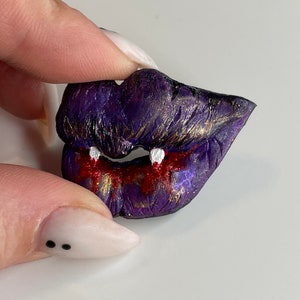 K&ouml;nnte beinhalten: Eine handgefertigte Brosche aus Polymer Clay in Form einer Lippe mit Glitzer in Lila und Gold, wei&szlig;en Z&auml;hnen und rotem, tropfendem Blut.
