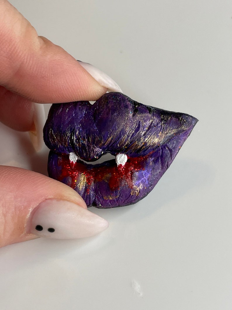 K&ouml;nnte beinhalten: Eine handgefertigte Brosche aus Polymer Clay in Form einer Lippe mit Glitzer in Lila und Gold, wei&szlig;en Z&auml;hnen und rotem, tropfendem Blut.