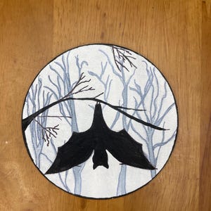 Pode incluir: Ilustra&ccedil;&atilde;o circular de um morcego preto pendurado em um galho. O fundo mostra uma cena de floresta com &aacute;rvores azul claro sobre um fundo branco. A obra de arte est&aacute; em uma superf&iacute;cie de madeira.