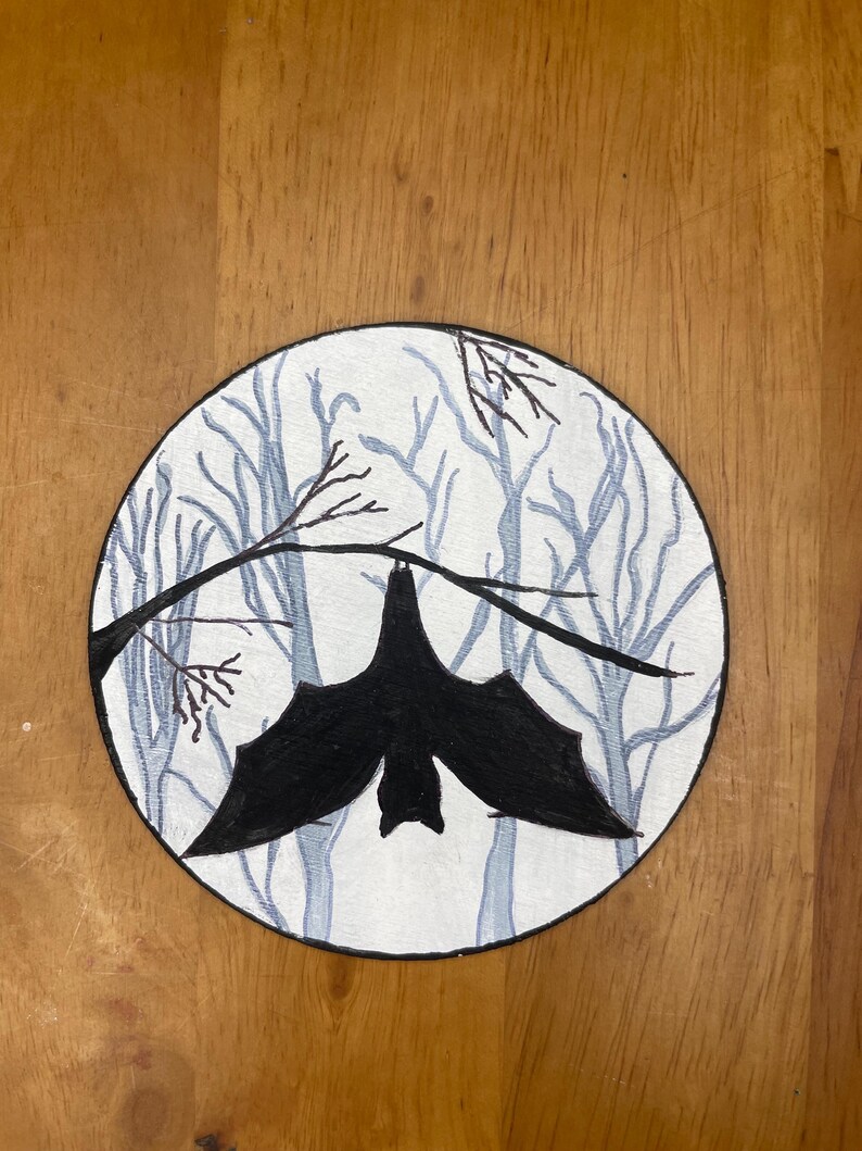 Pode incluir: Ilustra&ccedil;&atilde;o circular de um morcego preto pendurado em um galho. O fundo mostra uma cena de floresta com &aacute;rvores azul claro sobre um fundo branco. A obra de arte est&aacute; em uma superf&iacute;cie de madeira.