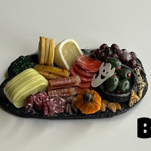 Op de afbeelding: Een zwart leisteen serveerbord met een Halloween-thema charcuterie arrangement. Het bord bevat verschillende kazen, vlees, olijven, druiven en een kleine pompoen. Een spookvormige koekjesuitsteker is op het bord geplaatst.