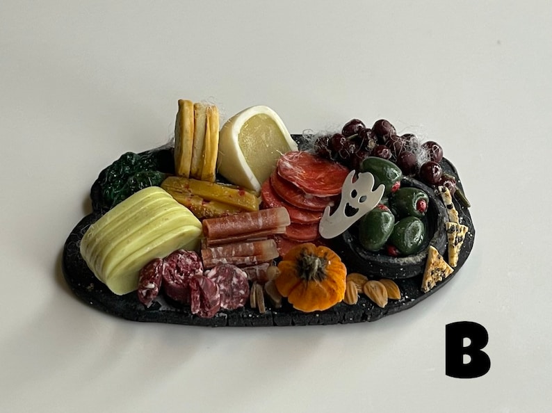 Op de afbeelding: Een zwart leisteen serveerbord met een Halloween-thema charcuterie arrangement. Het bord bevat verschillende kazen, vlees, olijven, druiven en een kleine pompoen. Een spookvormige koekjesuitsteker is op het bord geplaatst.