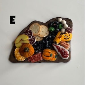 Op de afbeelding: Een miniatuur charcuterie-bord met een verscheidenheid aan herfstthema-voedingsmiddelen, waaronder crackers, salami, kaas, druiven, pompoenen en een schijf fruit. Het bord is gemaakt van donkerbruin hout en heeft een rustieke uitstraling.