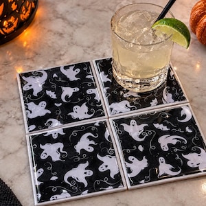 May include: A set of four square coasters featuring a repeating pattern of white ghosts on a black background. The coasters are arranged in a two-by-two grid, with a clear glass of liquid containing ice and a lime wedge resting on the top right coaster.