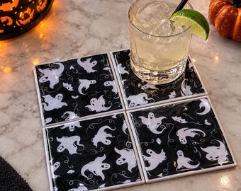 Puede incluir: Un juego de cuatro posavasos cuadrados con un patrón repetitivo de fantasmas blancos sobre un fondo negro. Los posavasos están dispuestos en una cuadrícula de dos por dos, con un vaso de cristal con hielo y una rodaja de lima encima.
