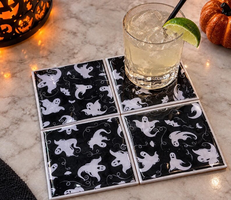 May include: A set of four square coasters featuring a repeating pattern of white ghosts on a black background. The coasters are arranged in a two-by-two grid, with a clear glass of liquid containing ice and a lime wedge resting on the top right coaster.