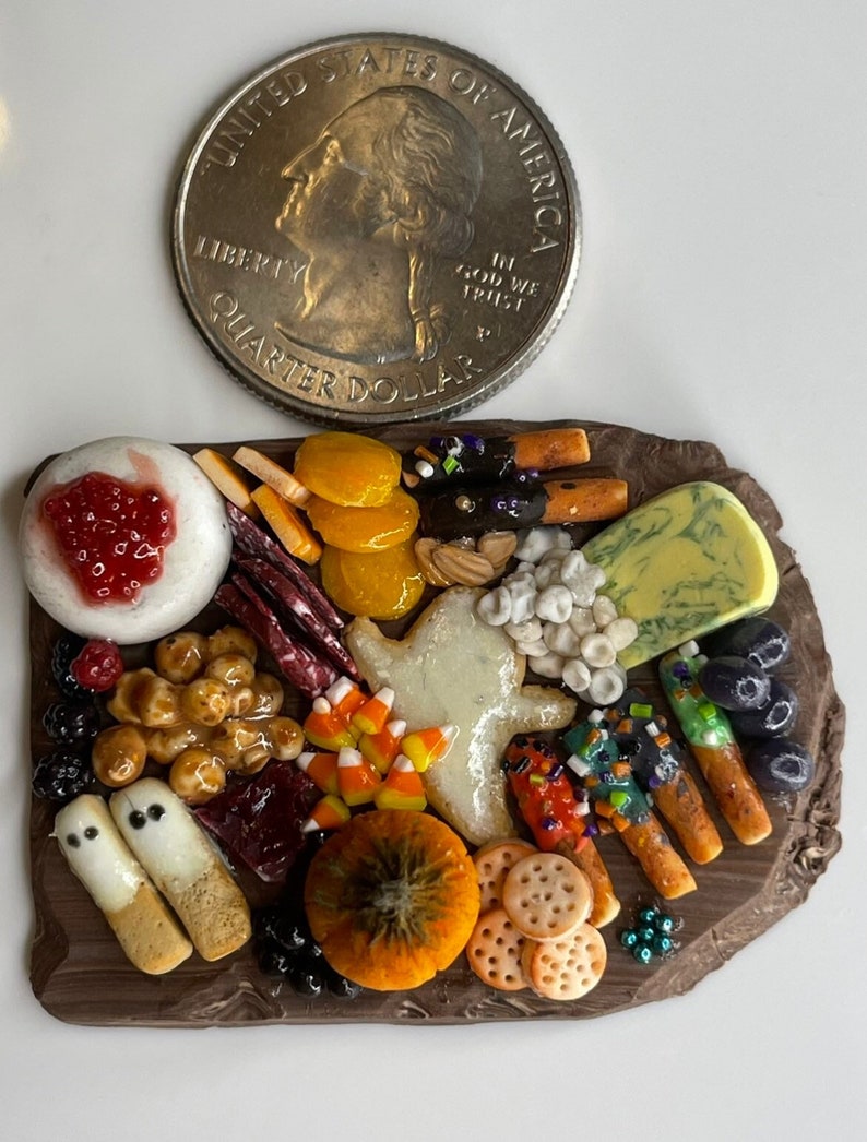 Op de afbeelding: Een miniatuur Halloween-charcuteriebord met een verscheidenheid aan snacks, waaronder kaas, crackers, fruit, candy corn en koekjes in de vorm van een spook. Het bord is gemaakt van bruine klei en is ongeveer zo groot als een kwartje.