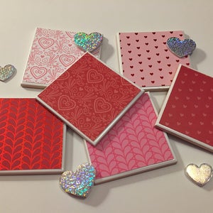 May include: Six square ceramic coasters with different heart patterns in shades of pink and red. The coasters have a white border. There are five iridescent heart-shaped decorations on the white surface.