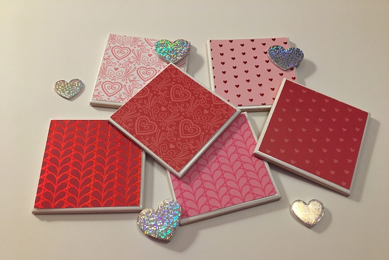 May include: Six square ceramic coasters with different heart patterns in shades of pink and red. The coasters have a white border. There are five iridescent heart-shaped decorations on the white surface.