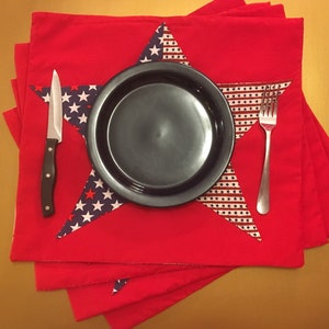 Peut inclure: Des sets de table rouges avec un motif en forme d'&eacute;toile repr&eacute;sentant un drapeau am&eacute;ricain bleu et blanc. Une assiette noire, une fourchette argent&eacute;e et un couteau argent&eacute; sont plac&eacute;s sur les sets de table.