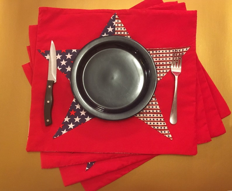 Peut inclure: Des sets de table rouges avec un motif en forme d'&eacute;toile repr&eacute;sentant un drapeau am&eacute;ricain bleu et blanc. Une assiette noire, une fourchette argent&eacute;e et un couteau argent&eacute; sont plac&eacute;s sur les sets de table.
