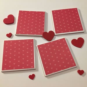May include: Four square ceramic coasters with a white border and a red background with a pattern of small white hearts. There are six red foam hearts of various sizes scattered around the coasters.