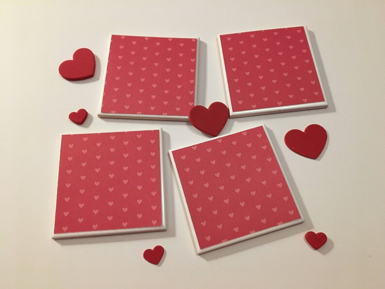 May include: Four square ceramic coasters with a white border and a red background with a pattern of small white hearts. There are six red foam hearts of various sizes scattered around the coasters.