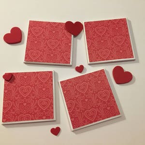 May include: Five red and white ceramic coasters with a heart pattern. Each coaster has a small red heart attached to the top.
