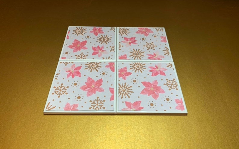 Peut inclure: Ensemble de quatre sous-verres en c&eacute;ramique blanche avec un motif de poinsettia rose et de flocons de neige dor&eacute;s.
