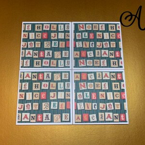 May include: Four square coasters with a Christmas theme. Each coaster features a green background with a white grid pattern. The coasters are decorated with red, white, and blue letters that spell out the words "HOLLY", "NICE", "JOY", "SANTA", "NOEL", "GLEN", "EVE", "ACE", and "SANT".