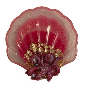 Puede incluir: Un plato en forma de concha rosa con un acabado brillante, que presenta un pulpo morado y rojo con detalles dorados y adornos perlados.