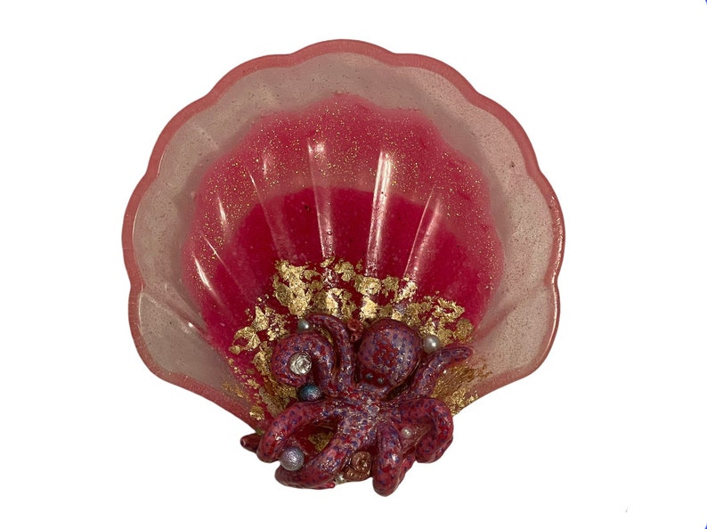 Puede incluir: Un plato en forma de concha rosa con un acabado brillante, que presenta un pulpo morado y rojo con detalles dorados y adornos perlados.
