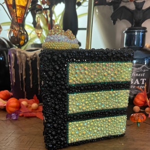May include: A black and green layered cake with a white and iridescent topper. The cake is covered in rhinestones and has a green border around each layer.