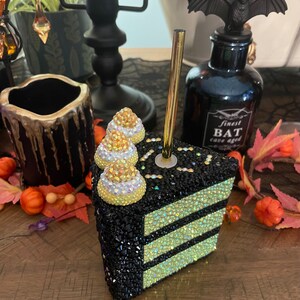 May include: A black and yellow cake slice with candy corn on top. The cake is covered in rhinestones and has a gold straw sticking out of the top.