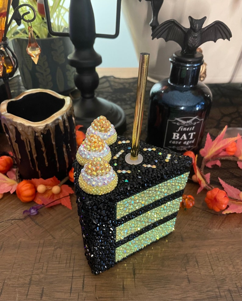 May include: A black and yellow cake slice with candy corn on top. The cake is covered in rhinestones and has a gold straw sticking out of the top.