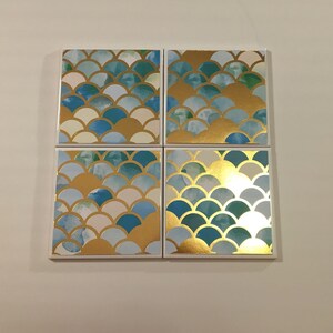 K&ouml;nnte beinhalten: Set mit vier quadratischen Untersetzern mit einem blauen und goldenen Schuppenmuster. Die Untersetzer sind aus wei&szlig;em Material und haben eine gl&auml;nzende Oberfl&auml;che.