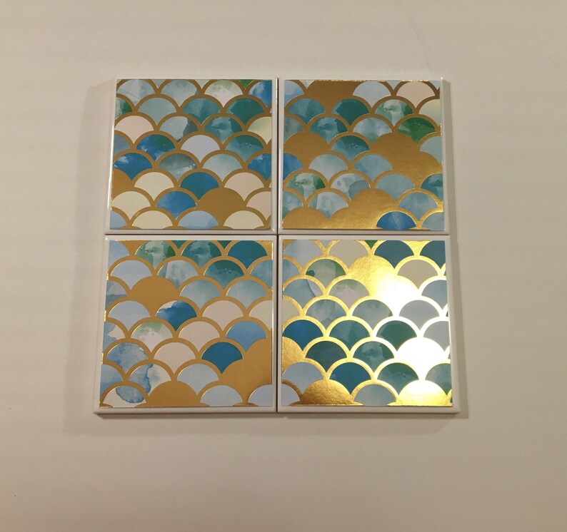 K&ouml;nnte beinhalten: Set mit vier quadratischen Untersetzern mit einem blauen und goldenen Schuppenmuster. Die Untersetzer sind aus wei&szlig;em Material und haben eine gl&auml;nzende Oberfl&auml;che.