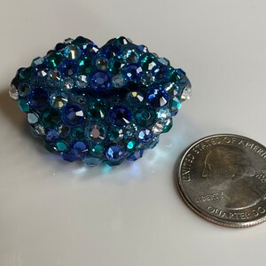 May include: A blue, jewel-encrusted lip-shaped object with varying shades of blue and clear crystals. A U.S. quarter coin is next to the object for size comparison. The object is likely a decorative item or accessory.