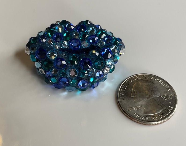 May include: A blue, jewel-encrusted lip-shaped object with varying shades of blue and clear crystals. A U.S. quarter coin is next to the object for size comparison. The object is likely a decorative item or accessory.