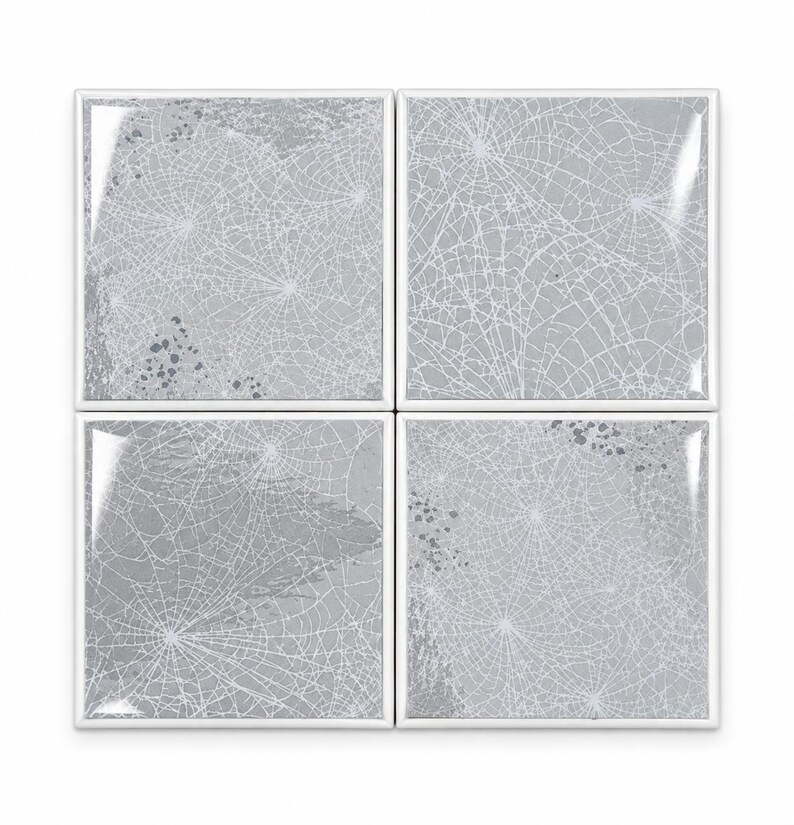 Peut inclure: Un ensemble de quatre carreaux de c&eacute;ramique carr&eacute;s dispos&eacute;s en grille, chacun pr&eacute;sentant une surface gris clair brillante avec un motif de toile d'araign&eacute;e blanc et des &eacute;claboussures de peinture gris fonc&eacute;, bord&eacute;s d'un contour blanc net.