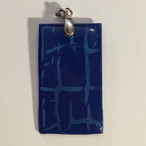 Puede incluir: Un colgante rectangular azul con una anilla de plata. El colgante tiene una superficie texturizada que se asemeja a un patr&oacute;n de ladrillo.