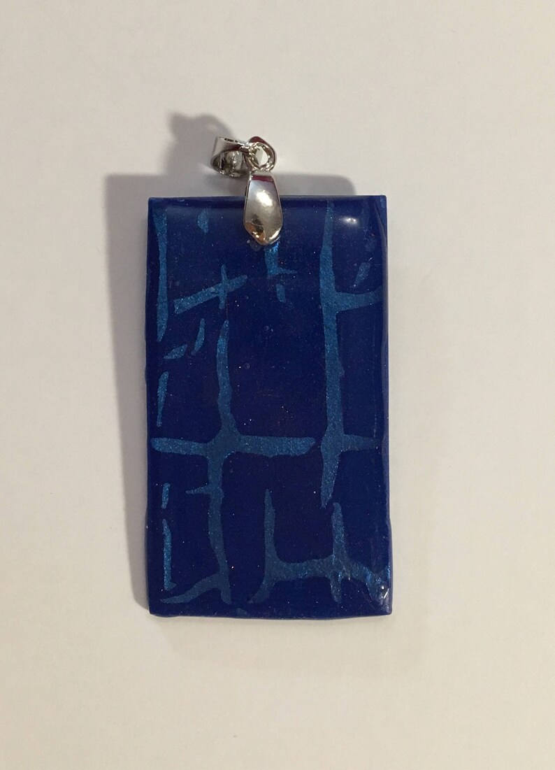 Puede incluir: Un colgante rectangular azul con una anilla de plata. El colgante tiene una superficie texturizada que se asemeja a un patr&oacute;n de ladrillo.