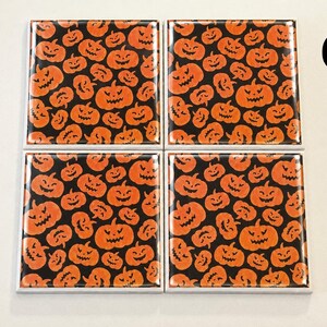 May include: A set of four square coasters featuring a repeating pattern of orange jack-o'-lanterns with carved faces on a black background. Each coaster has a glossy finish and white edges. The letter C is printed in black in the upper right corner.