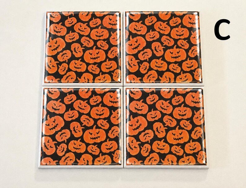 May include: A set of four square coasters featuring a repeating pattern of orange jack-o'-lanterns with carved faces on a black background. Each coaster has a glossy finish and white edges. The letter C is printed in black in the upper right corner.
