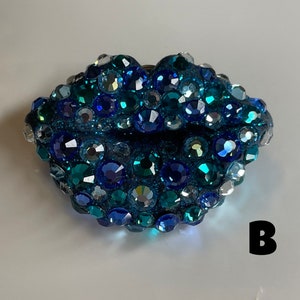 May include: A bedazzled, blue-toned lip-shaped brooch. The brooch is covered in various sizes of sparkling rhinestones in shades of blue, teal, and silver. The letter "B" is visible in the lower right corner.