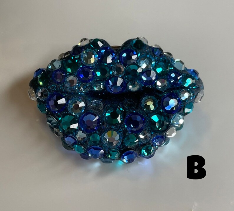 May include: A bedazzled, blue-toned lip-shaped brooch. The brooch is covered in various sizes of sparkling rhinestones in shades of blue, teal, and silver. The letter "B" is visible in the lower right corner.