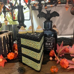 May include: A black and gold cake slice decorated with rhinestones and white frosting. The cake is decorated with a black bat and the text "finest BAT cave aged".