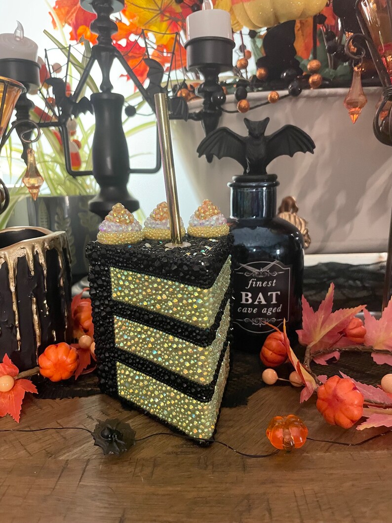 May include: A black and gold cake slice decorated with rhinestones and white frosting. The cake is decorated with a black bat and the text "finest BAT cave aged".