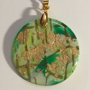 Puede incluir: Un colgante redondo verde, blanco y dorado con una anilla dorada. El colgante tiene un efecto marmoleado con detalles de pan de oro.