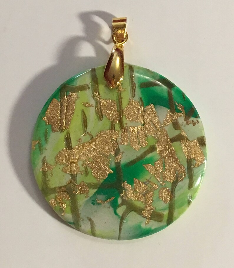 Puede incluir: Un colgante redondo verde, blanco y dorado con una anilla dorada. El colgante tiene un efecto marmoleado con detalles de pan de oro.